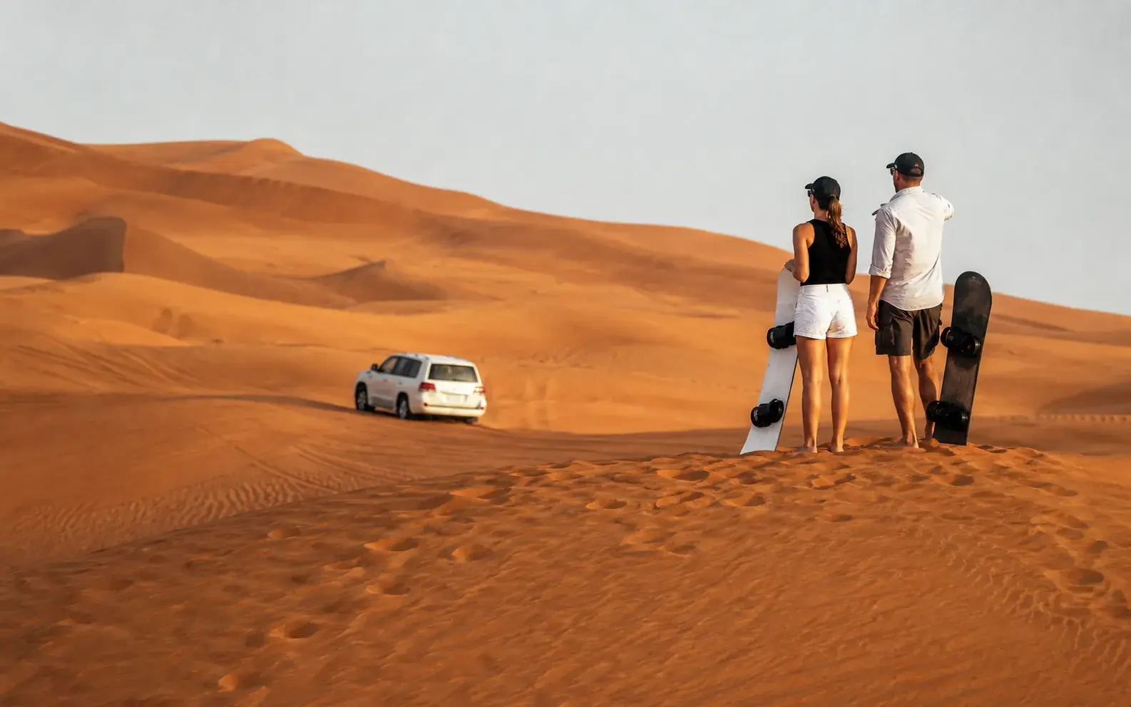 Safari vehicle crossing red dunes while travellers prepare sandboards on a nearby ridge