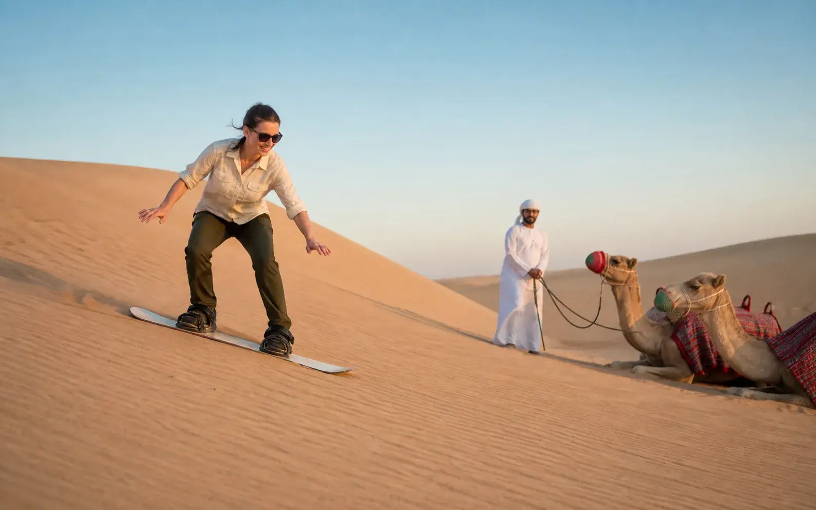 Traveller sandboarding beside a camel stop in clear morning light